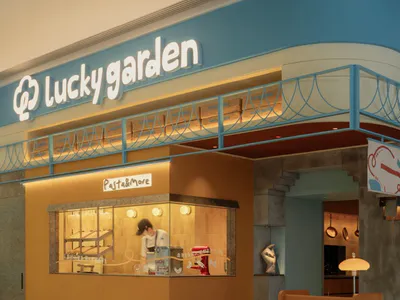 Modern commercial pasta and more kiosk named Lucky Garden with warm orange wood paneling, blue curved ceiling, illuminated display window showing a chef and red stand mixer, and a wooden host stand in the foreground.