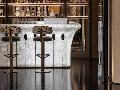 Luxurious modern home bar with a white and grey veined Calacatta marble curved counter, two patterned bar stools with dark green velvet seats, backlit dark metal and glass display shelves filled with liquor bottles and crystal glassware, and a sculptural alabaster wall sconce on the right.