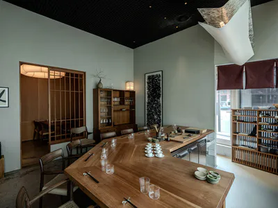 Modern Japanese restaurant interior with large L-shaped wooden dining counter, woven chairs, shoji-style sliding doors, and minimalist table settings with ceramic cups and chopsticks.
