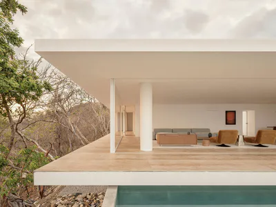 Modern open-plan living room with concrete overhang, large glass walls, mustard swivel chairs, and a sweeping view of a dry hillside and infinity pool.