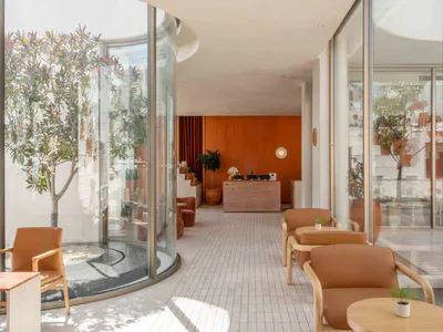 Modern hotel lobby interior with curved glass walls, terracotta reception desk, wooden armchairs with orange upholstery, and potted trees under bright natural light.