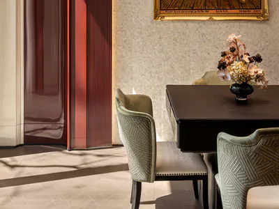 Elegant dining area with dark wood table, green patterned velvet chairs, a large ornate gold-framed surreal painting of a wooden floor with floating sticks, a modern circular chandelier, and a tall red lacquered wall panel with wall sconce.