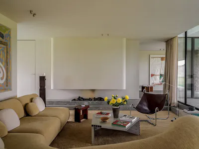 Modern minimalist living room featuring a curved beige sectional sofa, dark brown leather butterfly chair, metal and glass coffee table, and a recessed fireplace with large abstract paintings.