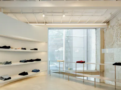 現代極簡服裝精品店室內空間，擁有大面積清水模牆面、白色懸浮展示層板、折疊衣物、不鏽鋼服裝展示架，以及落地窗引入的明亮自然光。