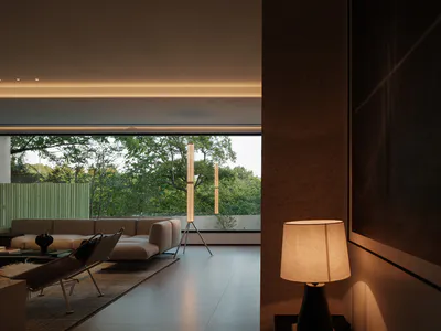 Modern minimalist living room featuring raw concrete walls, large floor-to-ceiling windows overlooking a green garden, a beige sectional sofa, a woven lounge chair, and warm ambient lighting from a tripod floor lamp and a table lamp.