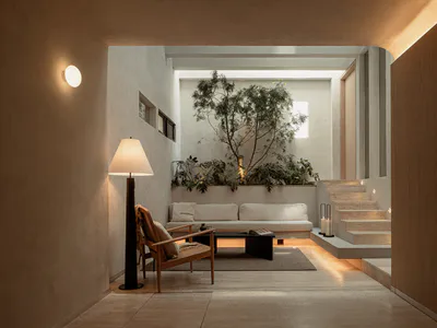 Modern minimalist living room featuring raw concrete walls, a wooden armchair, white sofa, tall floor lamp, and a large indoor tree in a raised planter, with a terrazzo staircase on the right.