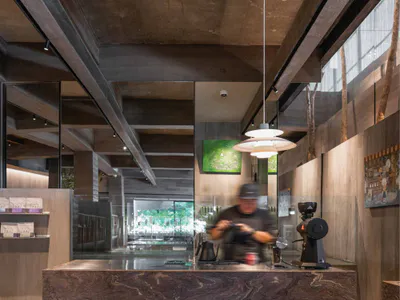 Modern industrial-style coffee shop interior featuring a large dark marble service counter, concrete ceiling, wooden walls, and a barista working behind the counter with coffee equipment.