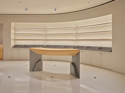 Modern minimalist retail interior featuring a curved light oak reception desk with angular concrete base, set against a curved wall with illuminated floating shelves and white glossy floor.