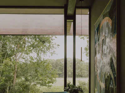Modern living room interior featuring a teal leather armchair, round wooden side table, large abstract painting, potted plant, and expansive floor-to-ceiling windows overlooking a green forest and meadow.