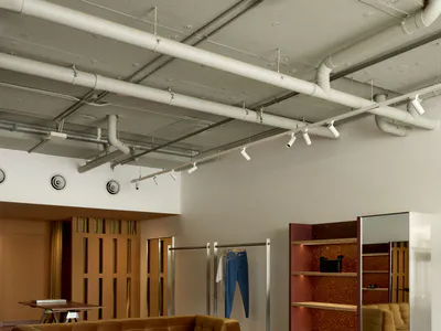 Industrial-style retail showroom with a brown tufted velvet sofa, metallic coffee table, red plastic chair, woven jute rug, and exposed white ceiling pipes.