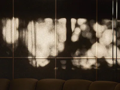 A curved brown boucle fabric modular sofa with a wooden side table holding a glass of iced matcha latte and a glass of water, set against a dark speckled wall with dramatic tree-like light and shadow patterns.