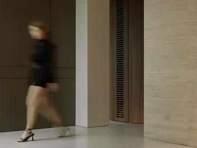 A person captured in motion blur walking through a modern minimalist interior hallway featuring a large travertine stone wall, dark wood cabinetry, and a wooden louvered door under soft ambient lighting.