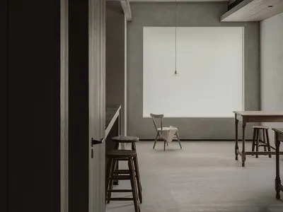 Minimalist open-plan dining and kitchen area featuring raw concrete walls and ceiling, wooden bar stools, a wooden dining table, a wooden chair with a draped cloth, and a single pendant light hanging in front of a large bright window.