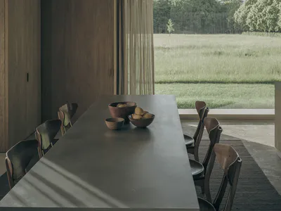 Modern minimalist dining room featuring a long concrete table, dark walnut wooden chairs, wooden cabinetry, and a large opening revealing a green meadow and forest beyond.