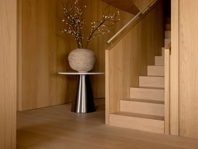 Modern minimalist entryway featuring a curved oak spiral staircase, light wood paneling, and a large stone vase with flowering branches on a conical metal pedestal.