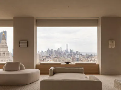Modern minimalist living room with beige boucle sofas and daybed, large floor-to-ceiling windows revealing the New York City skyline including One World Trade Center and the Woolworth Building under an overcast sky.