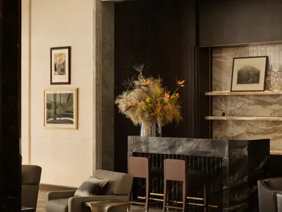 Elegant modern hotel lobby interior with dark marble reception desk, two wooden bar stools, a large dried floral arrangement with bird of paradise flowers, neutral upholstered armchairs on a patterned area rug, and framed artwork on light stone walls.