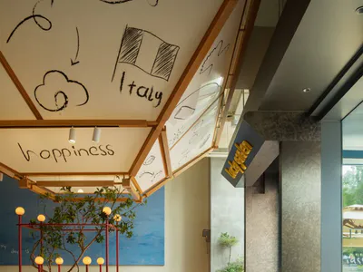Modern restaurant interior with wooden ceiling structure displaying hand-drawn mural of lucky, Italy flag, happiness, and cloud motifs, red bar stools, wooden tables and chairs, and a blue wall with potted tree.
