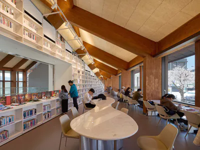 Modern library interior with tall white bookshelves, a large curved white marble island counter, wooden ceiling beams, and students studying at tables near large windows.