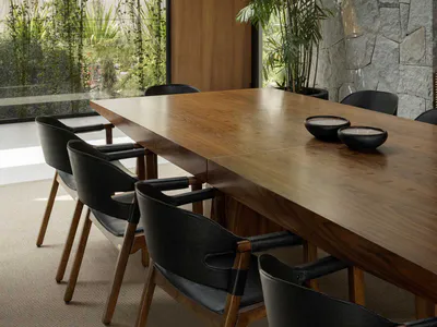 Modern dining room featuring a long walnut wood table, black leather dining chairs with wooden frames, a large circular black pendant light, a stacked stone wall, and expansive glass windows revealing lush hanging plants and greenery outside.