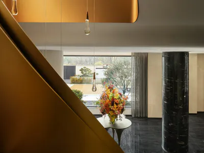 Modern double-height foyer featuring a curved golden brass wall, sculptural pendant lights, dark marble floor, and a large floral arrangement on a white table with a black marble column in the background.