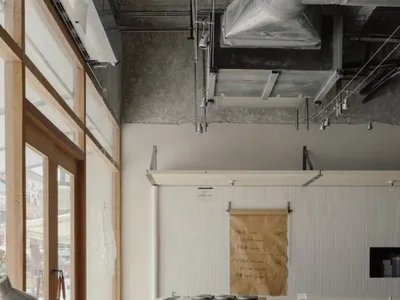 現代極簡咖啡廳廚房室內，包含淺木色櫃體、前景木製工作台、裸露混凝土天花板與管線，以及掛在白色直條板牆上的大型紙質菜單。