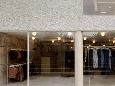 極簡精品服飾店外觀，粗糙灰泥牆面、大面積玻璃窗、室內可見服裝展示架與球形吊燈。