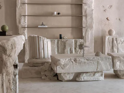 Minimalist living room featuring massive rough-hewn limestone blocks used as structural elements, coffee tables, and pedestals, with floating metal shelves mounted within a stone wall, decorative crown moldings on the upper wall, and neutral-toned accessories including a green sphere, dried botanicals, and sculptural objects.