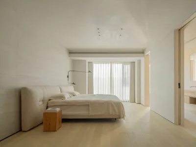 A serene minimalist bedroom with a low-profile upholstered bed, wooden nightstand, textured plaster walls, and large windows with sheer curtains allowing abundant natural light.