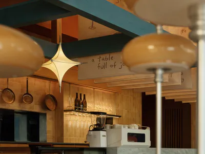 Interior view of a modern cafe bar featuring a prominent blue structural beam, starburst pendant light, hanging copper pans, and a service counter with a blender and display case.