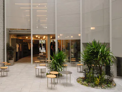 Modern minimalist courtyard between concrete buildings featuring sheer white fabric screens, round wooden stools, metal tables, and lush green plants.