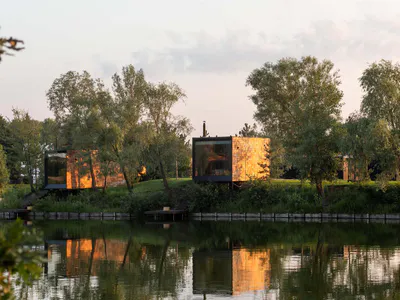 Two modern floating cabins with large glass windows and corten steel cladding situated beside a calm lake surrounded by lush green trees at sunset.