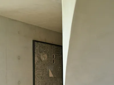 View of a curved concrete staircase and wall featuring a large framed artwork with intricate tropical etching and geometric border patterns against exposed concrete walls.