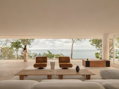 Modern minimalist living room with two burnt-orange swivel chairs, low teak coffee tables, and a woman in a white dress standing by the edge of an open terrace overlooking the ocean and trees.