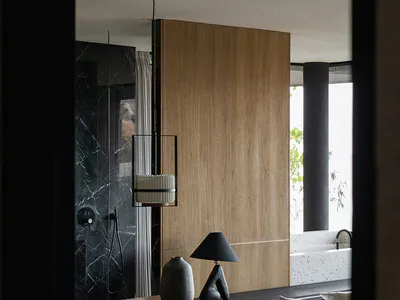 Modern bathroom interior viewed through a doorway, featuring a wooden vanity desk with a sculptural black lamp and ceramic vase, a wooden chair, a large warm wood panel wall, a black marble shower area, and a white terrazzo bathtub by a window.