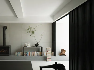 Modern minimalist living room with concrete walls, black fireplace, raised platform with books and bamboo plant, white cylindrical pendant lamp, and black dining chairs.