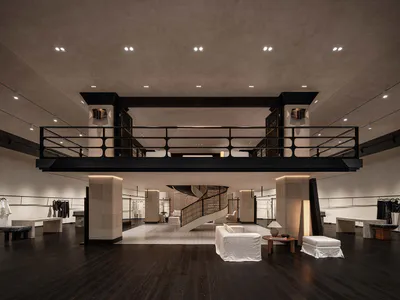 Spacious luxury boutique interior with double-height ceiling, black metal mezzanine railing, travertine stone columns, dark wood floor, white upholstered seating, and a curved black spiral staircase in the background.