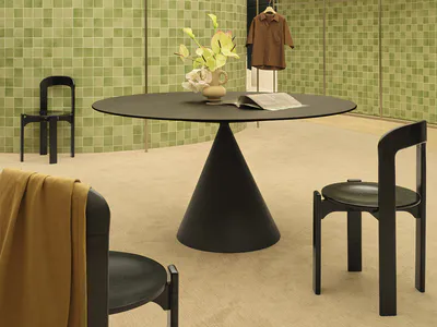 Modern minimalist boutique interior featuring curved walls covered in small green ceramic tiles, a large black conical-base round table, matching black wooden chairs, and a beige carpeted floor with soft natural lighting from a linear ceiling.