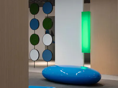 Modern corporate lounge interior with glossy blue organic-shaped seating pods, hanging circular moss and colored panels, wood slat ceiling, and a glowing green vertical light fixture.