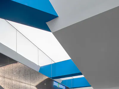 Contemporary tech company lounge with modular blue and grey sofas, dramatic angular blue ceiling panels, and large illuminated walls featuring intricate white circuit board patterns under a bright skylight.