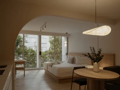 Modern minimalist bedroom featuring a low platform bed with wooden headboard, round light oak dining table with black chairs, dark sofa, large arched doorway, and floor-to-ceiling glass doors opening to a balcony with greenery.