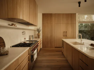 Modern minimalist kitchen featuring light oak cabinetry, white countertops, a central island with sink, and stainless steel range.