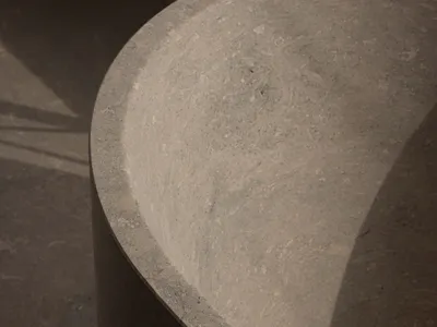 Close-up view of a thick-walled, handcrafted concrete or stone basin with a textured surface, set against a dark concrete floor with strong directional shadows.