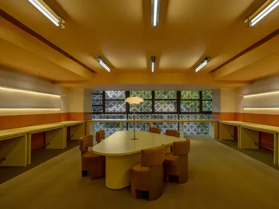 現代機構書房，中央為大型奶油色橢圓桌，周圍環繞六張焦橙色絨布旋轉椅，嵌入式淺木色周邊書桌搭配溫暖橙色飾板，大型圖案窗戶可眺望綠意。天花板設有線性日光燈，地毯為米色紋理。