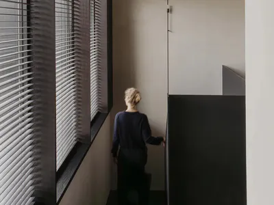 Woman with blonde hair in a bun walking up a modern concrete staircase next to a tall window with dark grey venetian blinds in a minimalist interior.