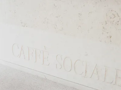 Close-up view of a white marble wall featuring raised, carved lettering that reads 'CAFFÈ SOCIALE' in an elegant serif font, with a speckled terrazzo floor visible below.