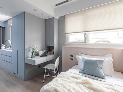 Modern bedroom interior with blue-grey built-in wardrobe and desk unit, white floating desk with laptop, light wood bed with blue and white bedding, and large window with roller blind.