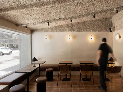Interior view of a modern minimalist cafe with a heavily textured plaster ceiling, terracotta floor tiles, wooden tables, built-in banquette seating, and a person walking through the space.
