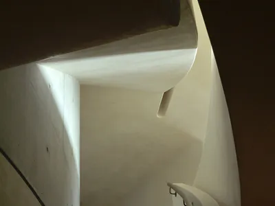 View looking down a curved concrete spiral staircase with a smooth handrail and dramatic natural light creating strong contrasts on the walls.