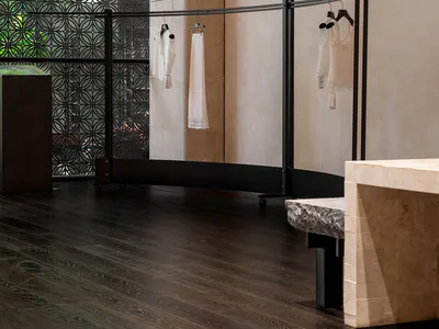 極簡精品店室內景觀，弧形黑色金屬服裝展示架上掛著白色蕾絲與刺繡服飾，前景有大型洞石展示座，深色木地板，背景為裝飾性綠色圖案窗屏。
